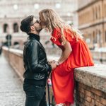 man standing and kissing woman sitting on concrete wall